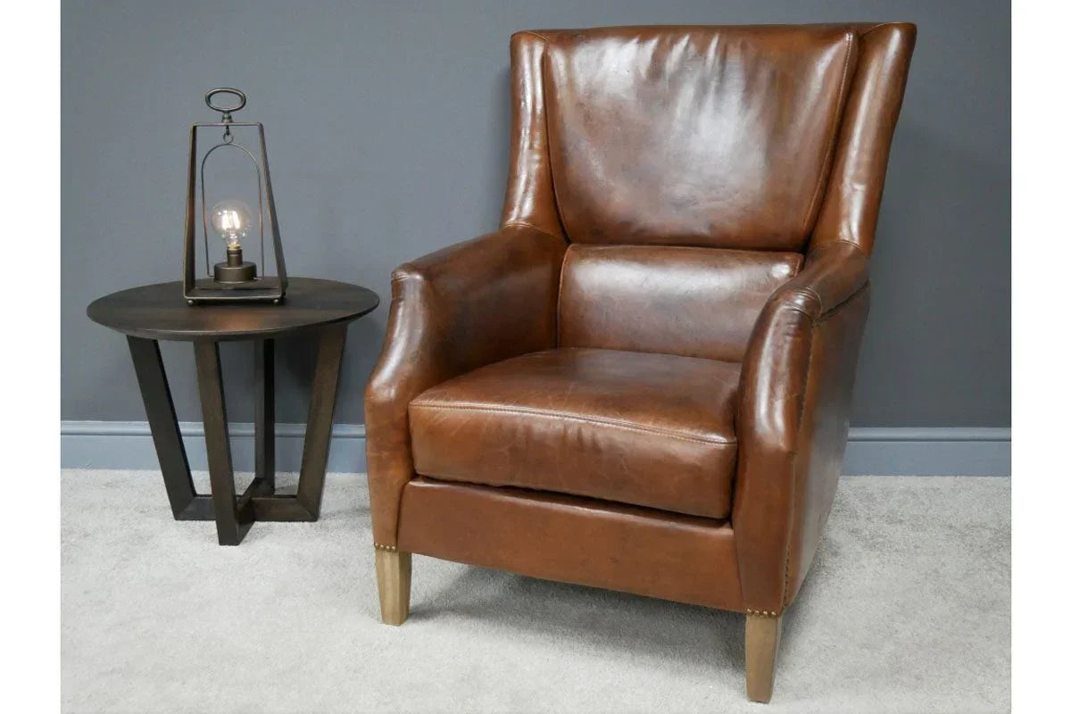 Brown leather armchair beside a round wooden side table with lamp on grey carpet