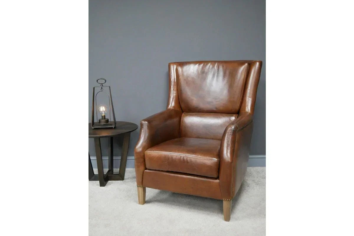 Brown leather armchair with high backrest next to round side table and lamp on grey carpet