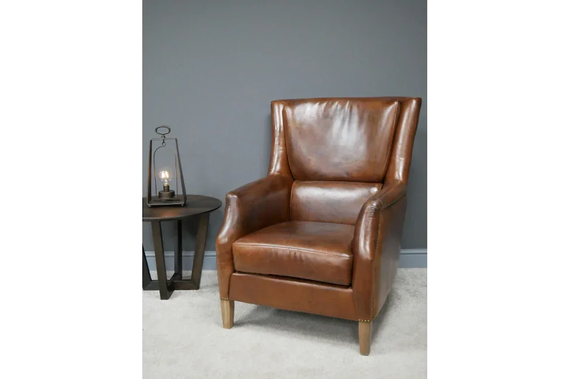 Brown leather armchair with high back next to round wooden side table and lamp