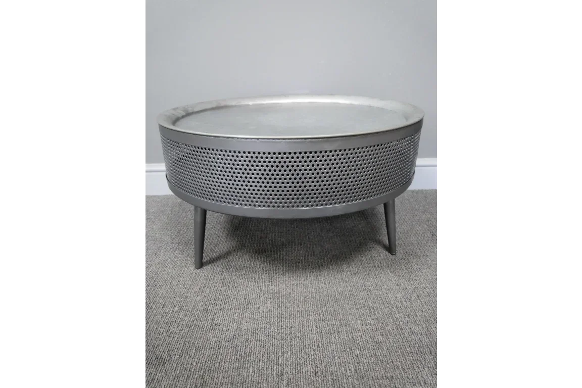 Round metal coffee table with perforated sides and tripod legs on grey carpet