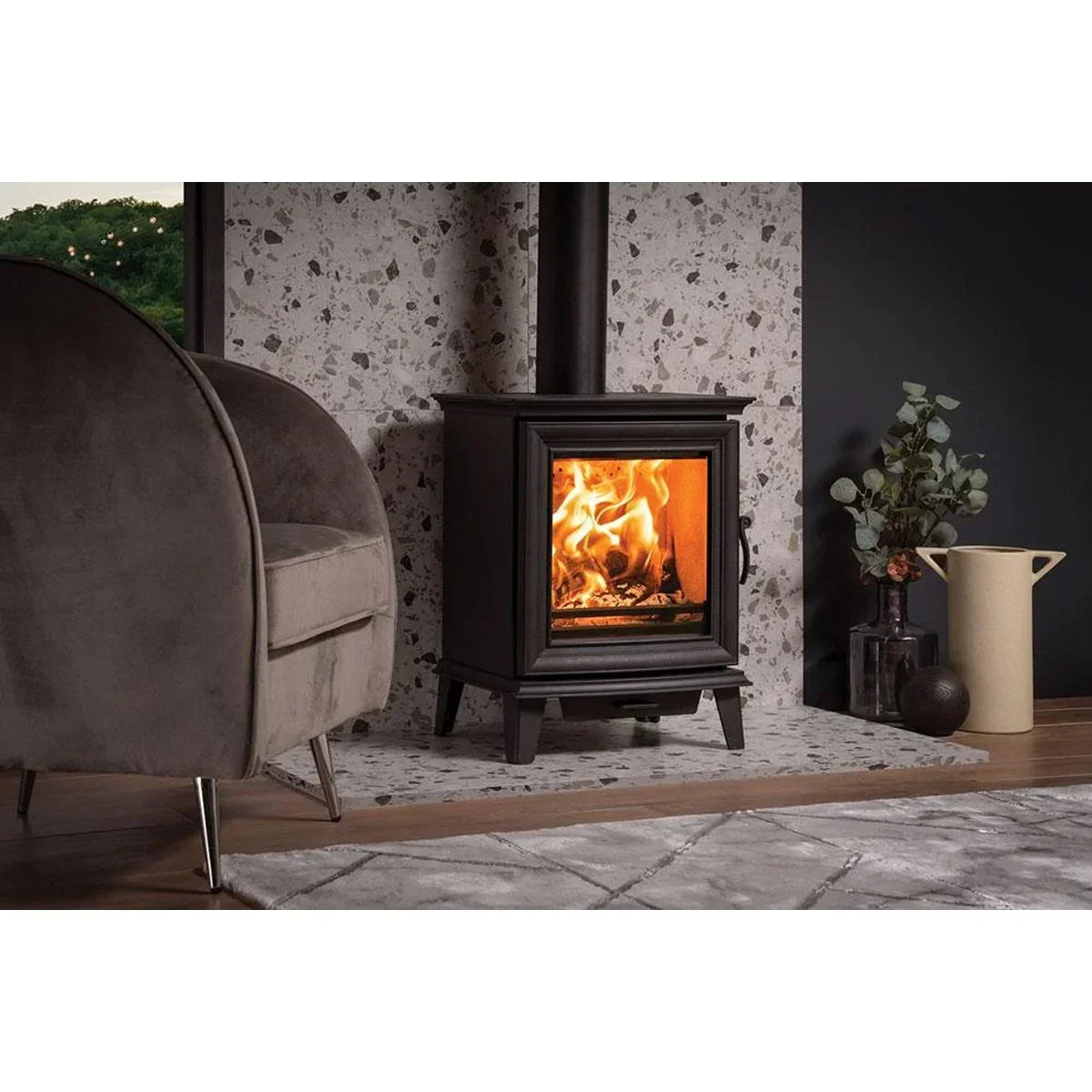 Modern black wood burning stove with flames beside armchair and decorative vase