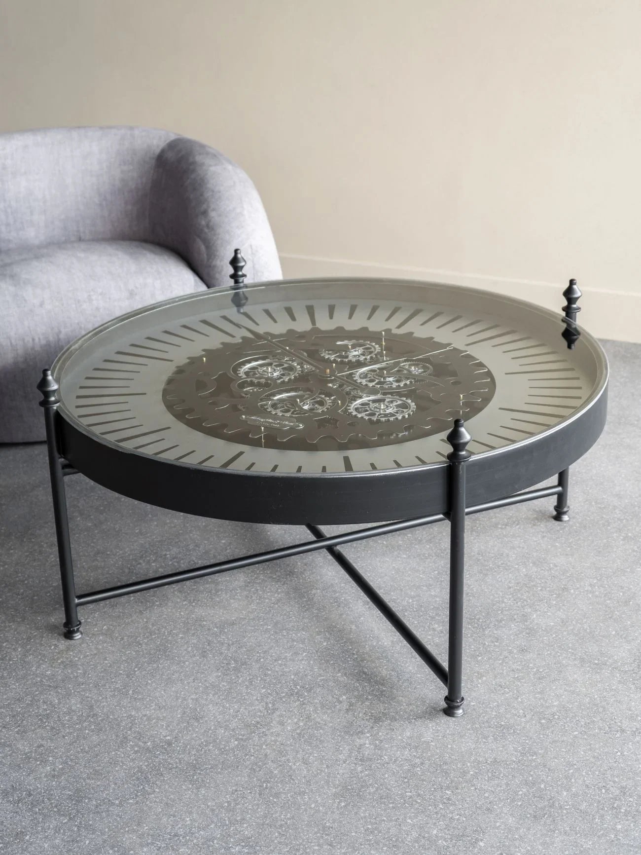 Round glass coffee table with visible cogs, set on a grey floor near a sofa.