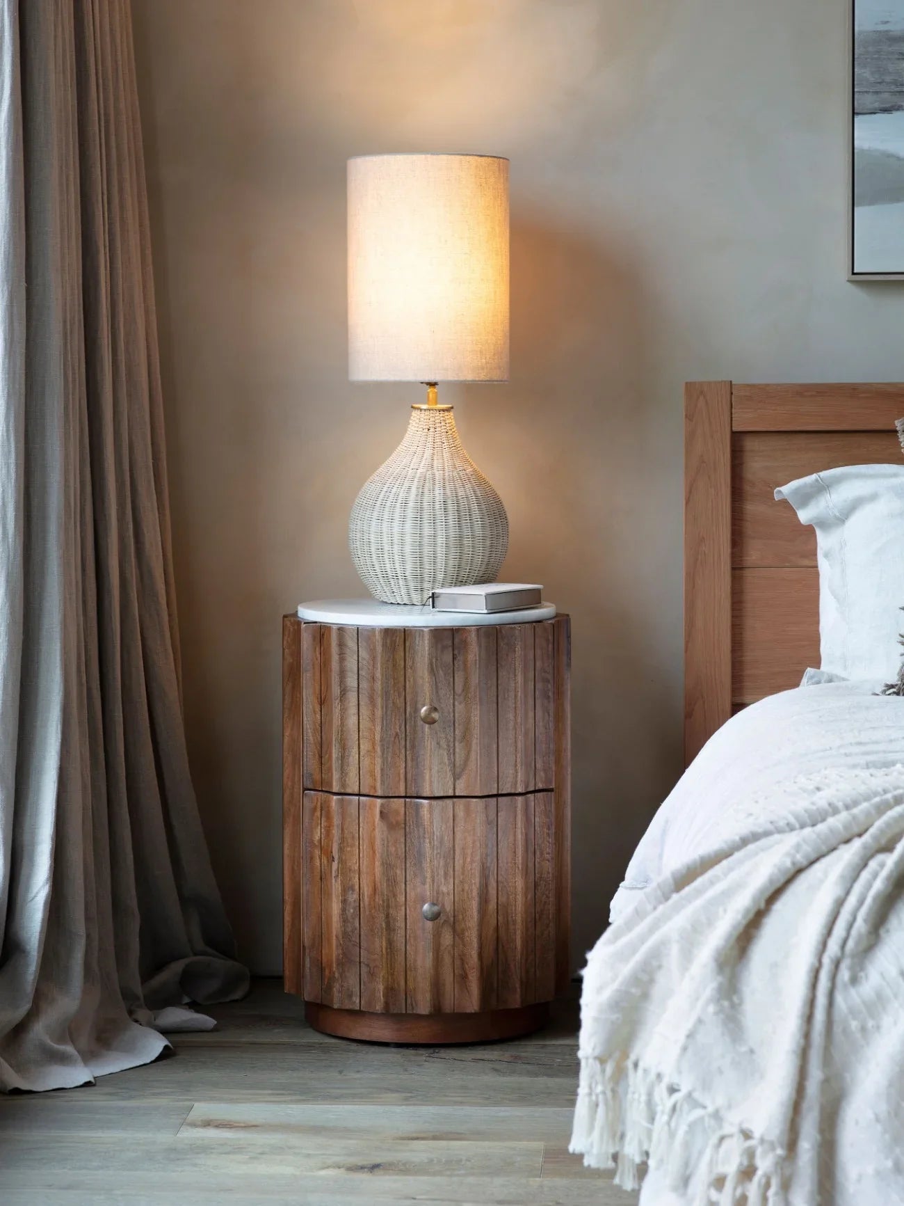 Wooden bedside table with marble top beside bed and lamp in cozy bedroom.