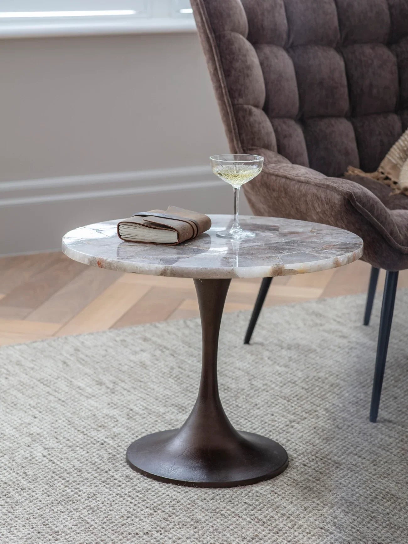 Round stone side table with a glass and book, next to a plush brown chair.