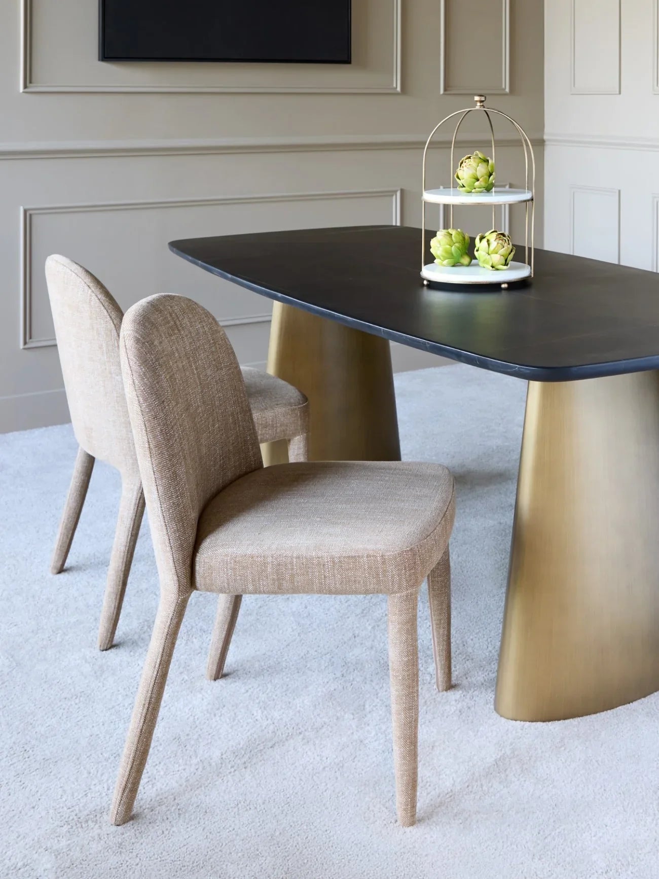 Wheat upholstered dining chairs beside a dark table with gold legs in a modern room.