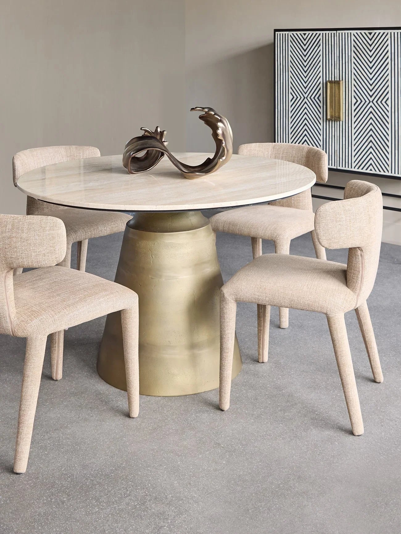 Beige dining chairs around a round travertine table with a bronze sculpture centerpiece.