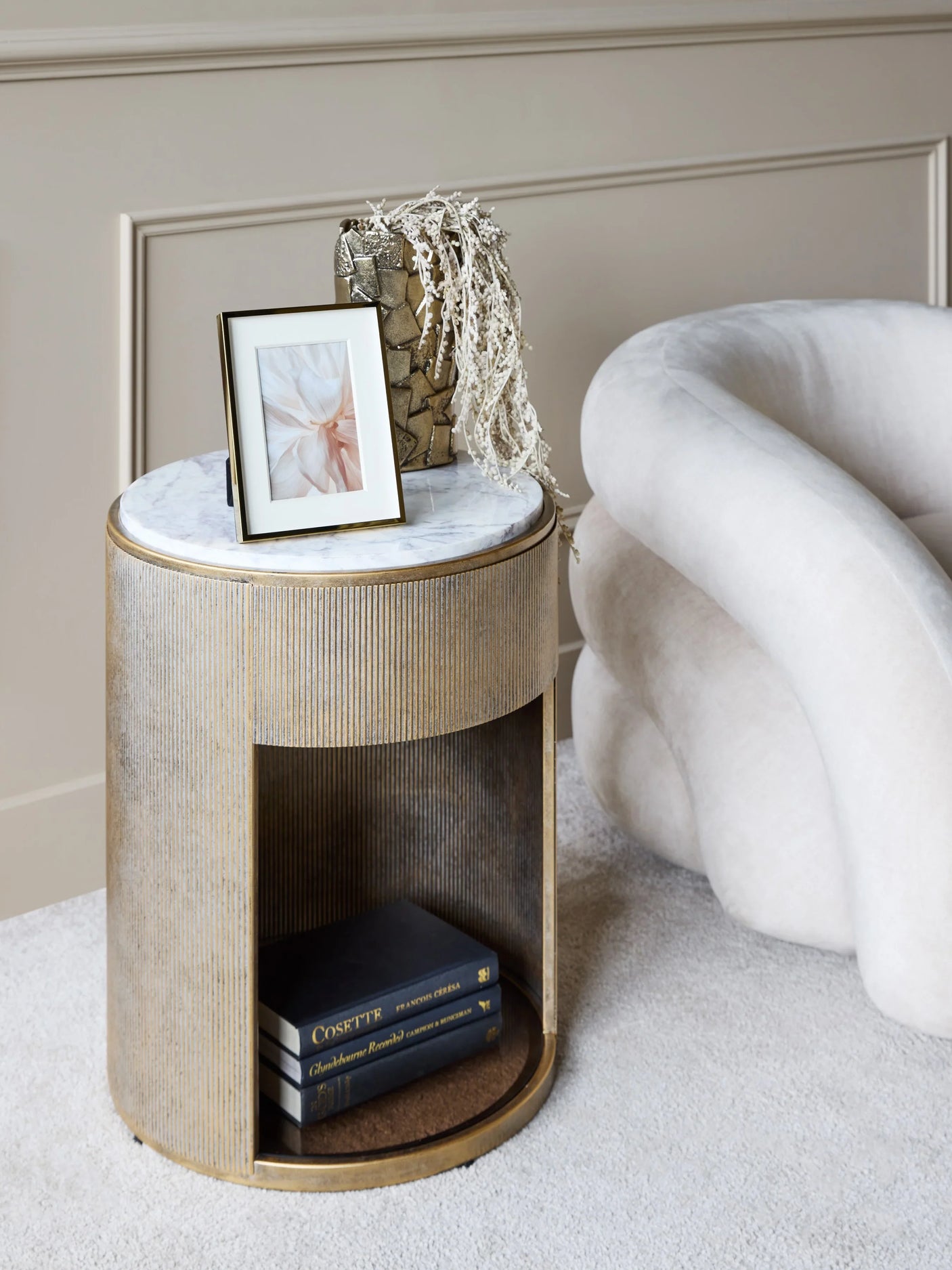 Aged gold side table with marble top next to a plush cream chair.