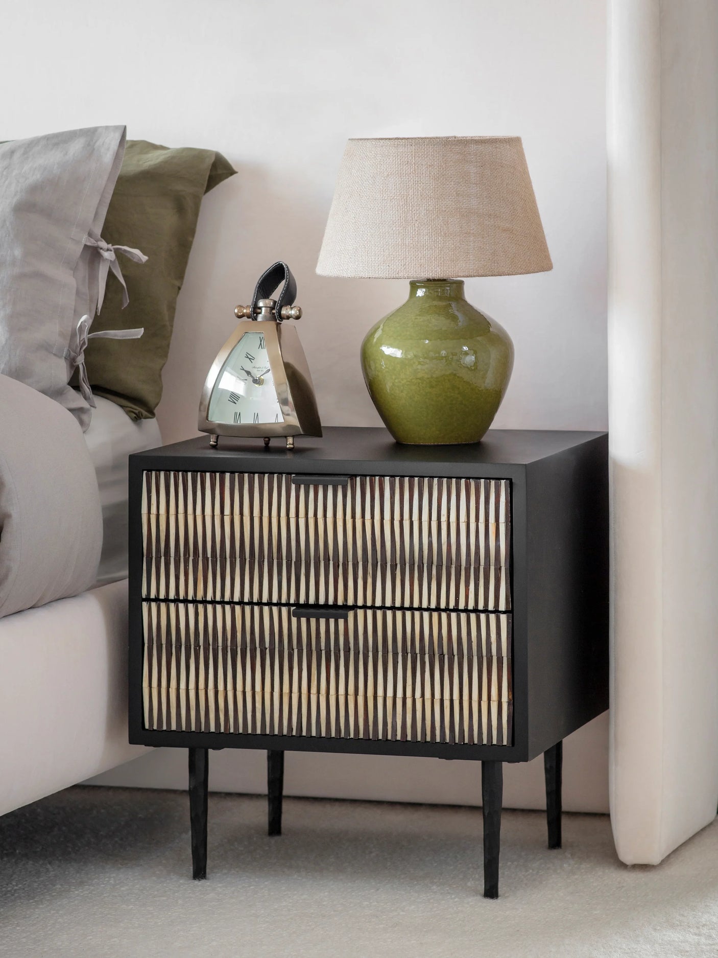 Black bedside table with bone inlay beside bed, topped with green lamp and clock.