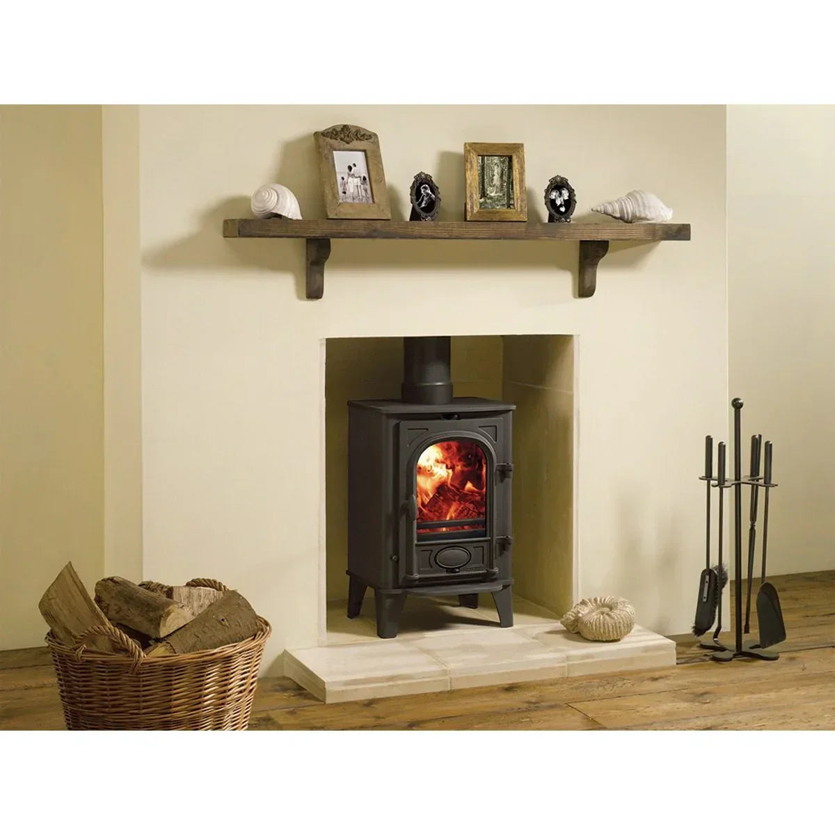 Black wood burning stove set in fireplace with basket of logs and fire tools nearby