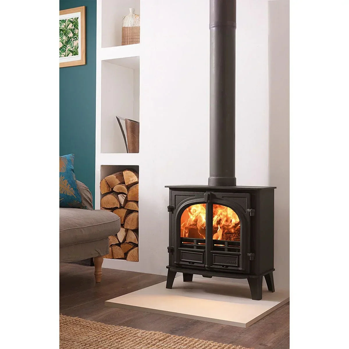 Black wood burning stove with fire, chimney pipe, and visible stacked logs in living room