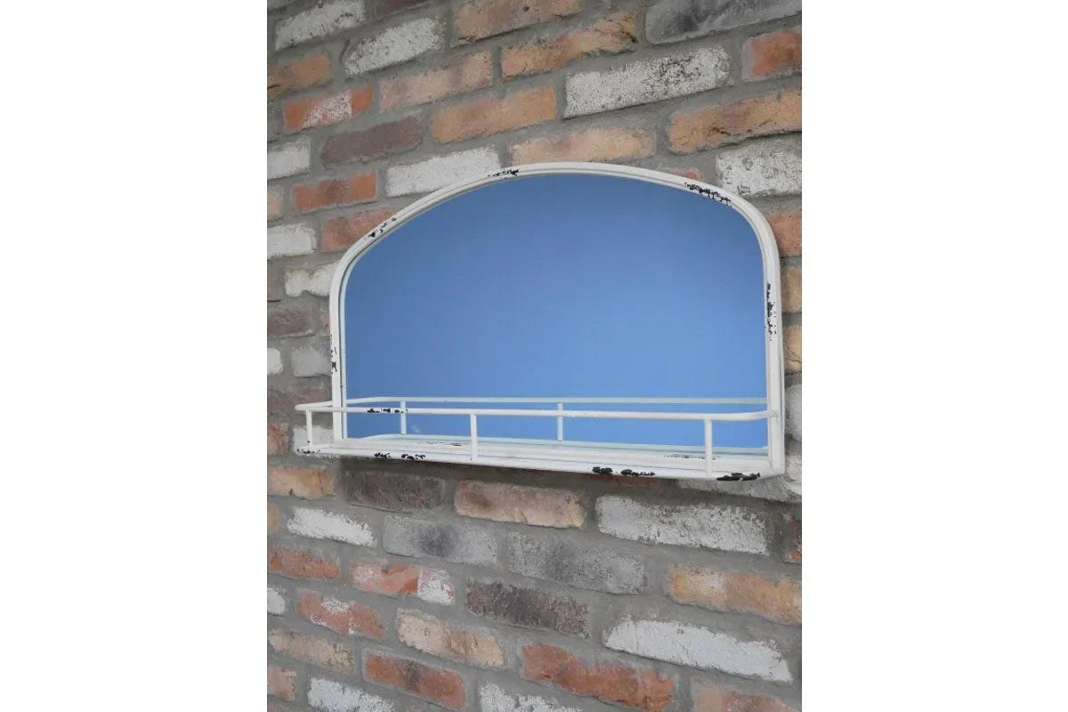 Arched wall mirror with distressed white metal frame and shelf mounted on brick wall