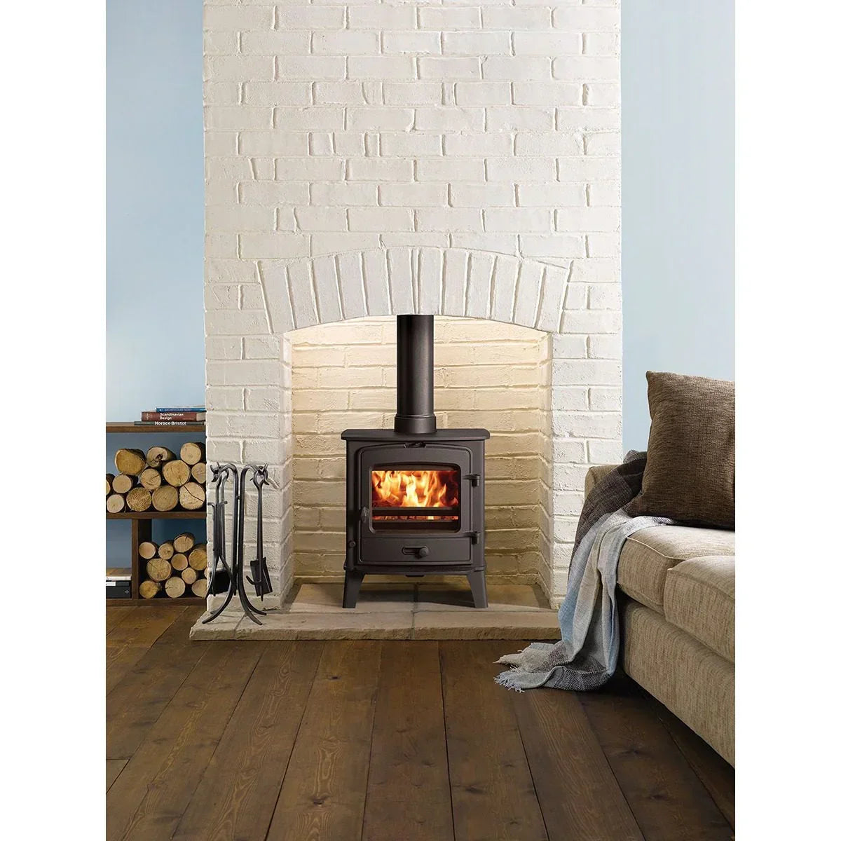 Black wood burning stove in white brick fireplace with log storage and beige sofa nearby