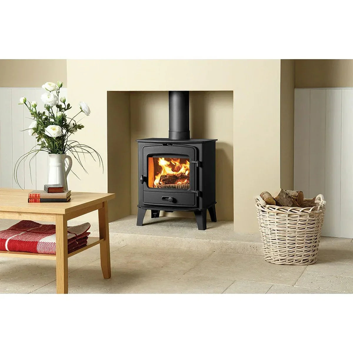 Black wood burning stove with fire, wicker log basket, coffee table and flower vase
