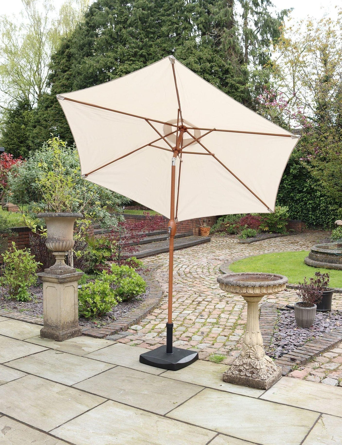 Cream garden parasol with wooden pole on stone patio beside birdbath and plants