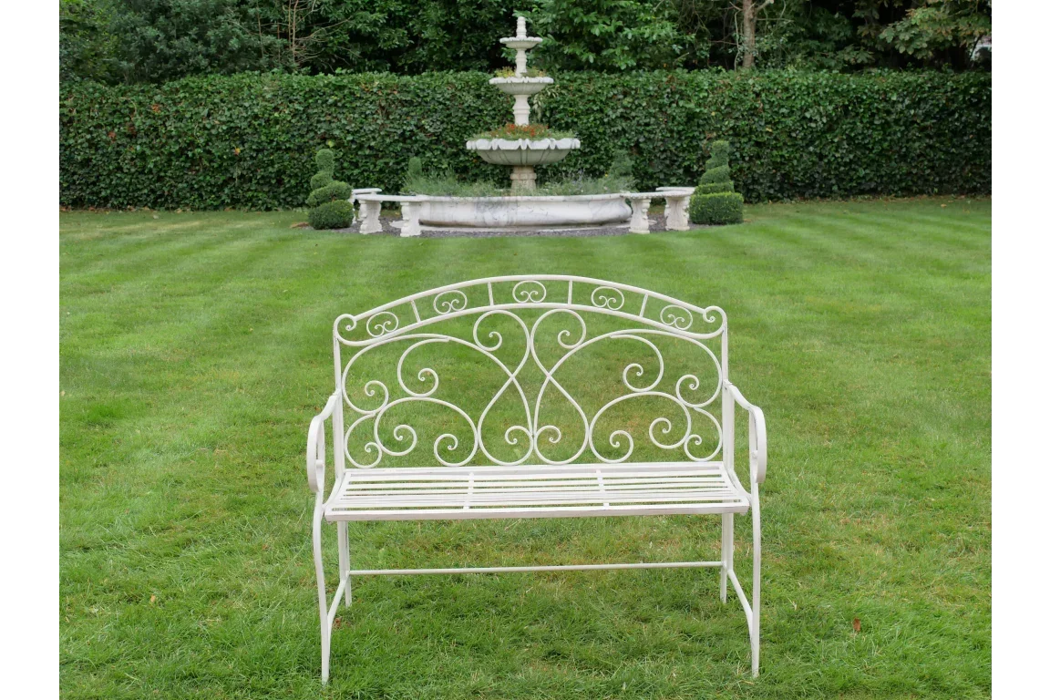 White metal garden bench with ornate scrollwork on lawn, fountain in background