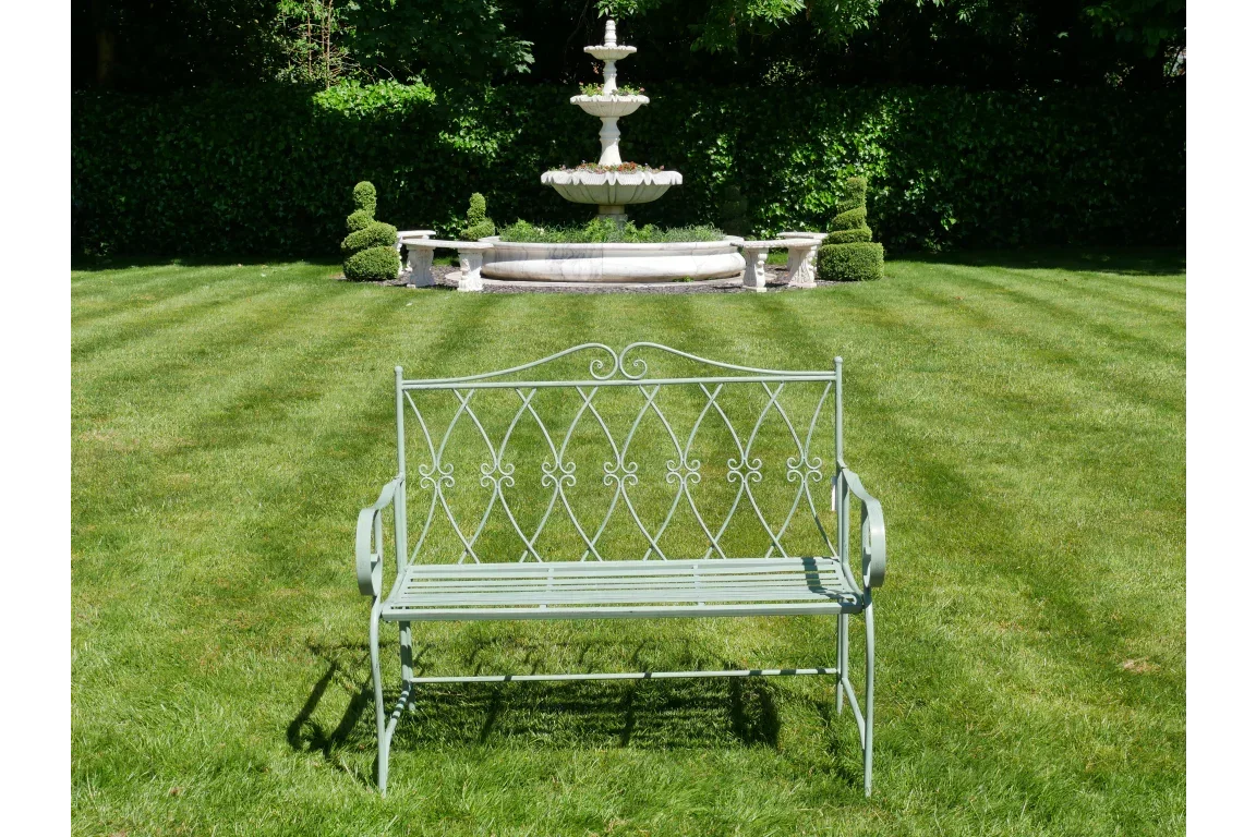 Metal garden bench with decorative backrest on grass, stone fountain in background