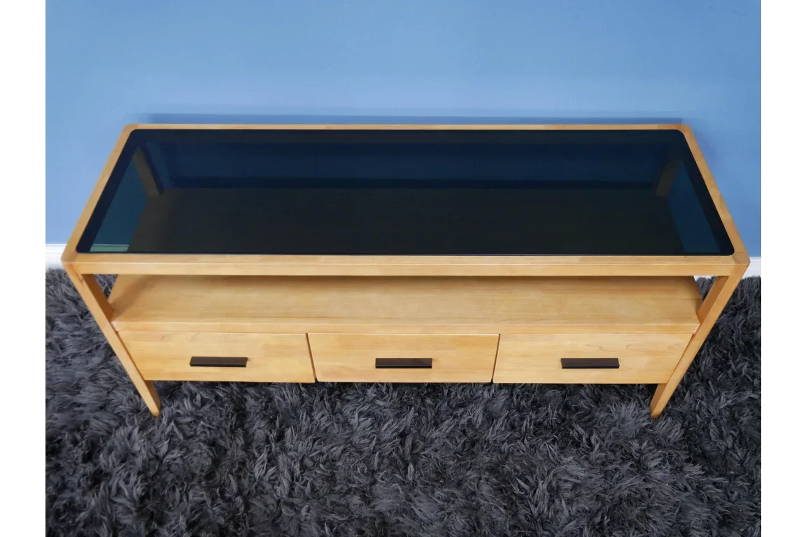 Oak TV unit with black glass top, three drawers and shelf on grey rug against blue wall