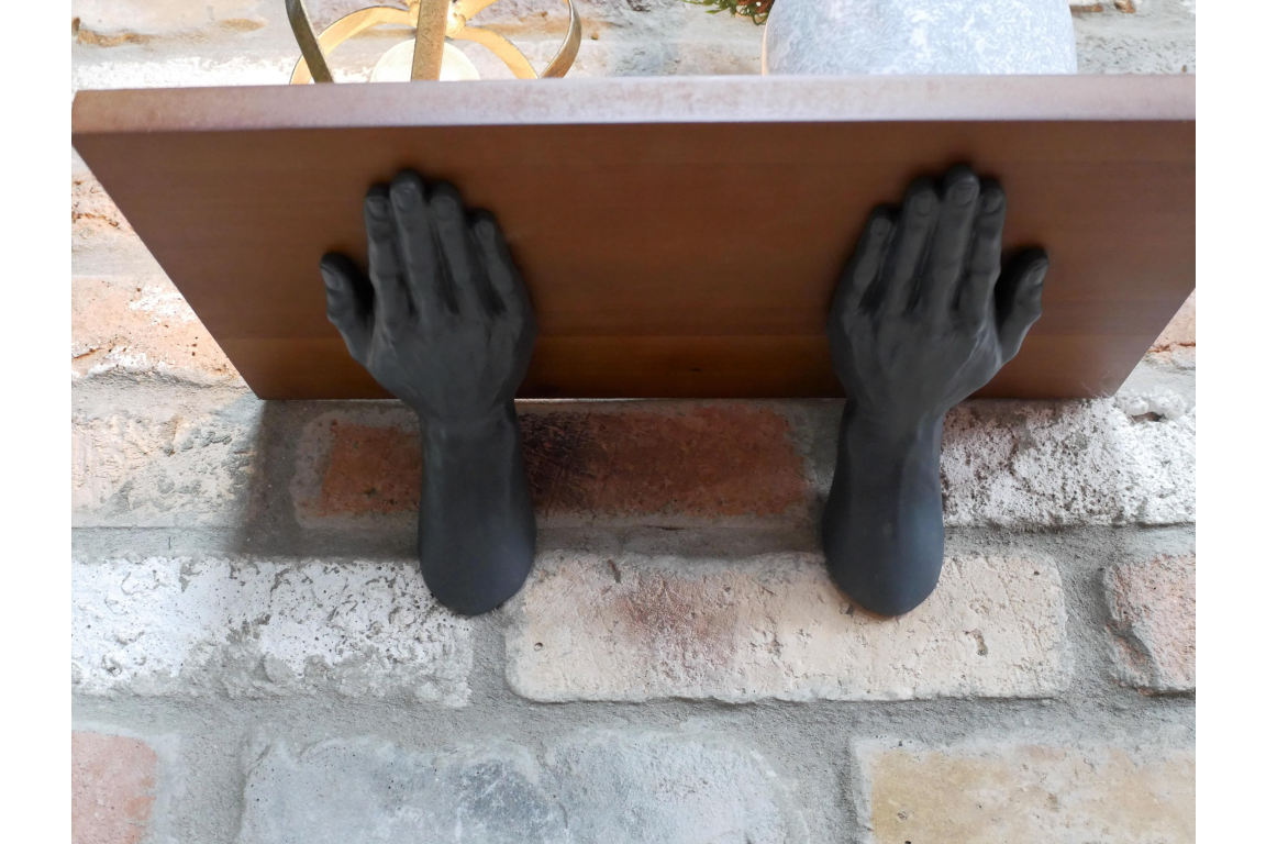 Wall-mounted wooden shelf supported by two black hand-shaped metal brackets against a brick wall.