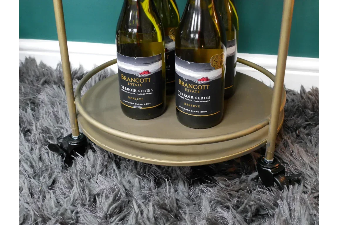 Gold round drinks trolley with wine bottles on lower shelf and swivel caster wheels on rug