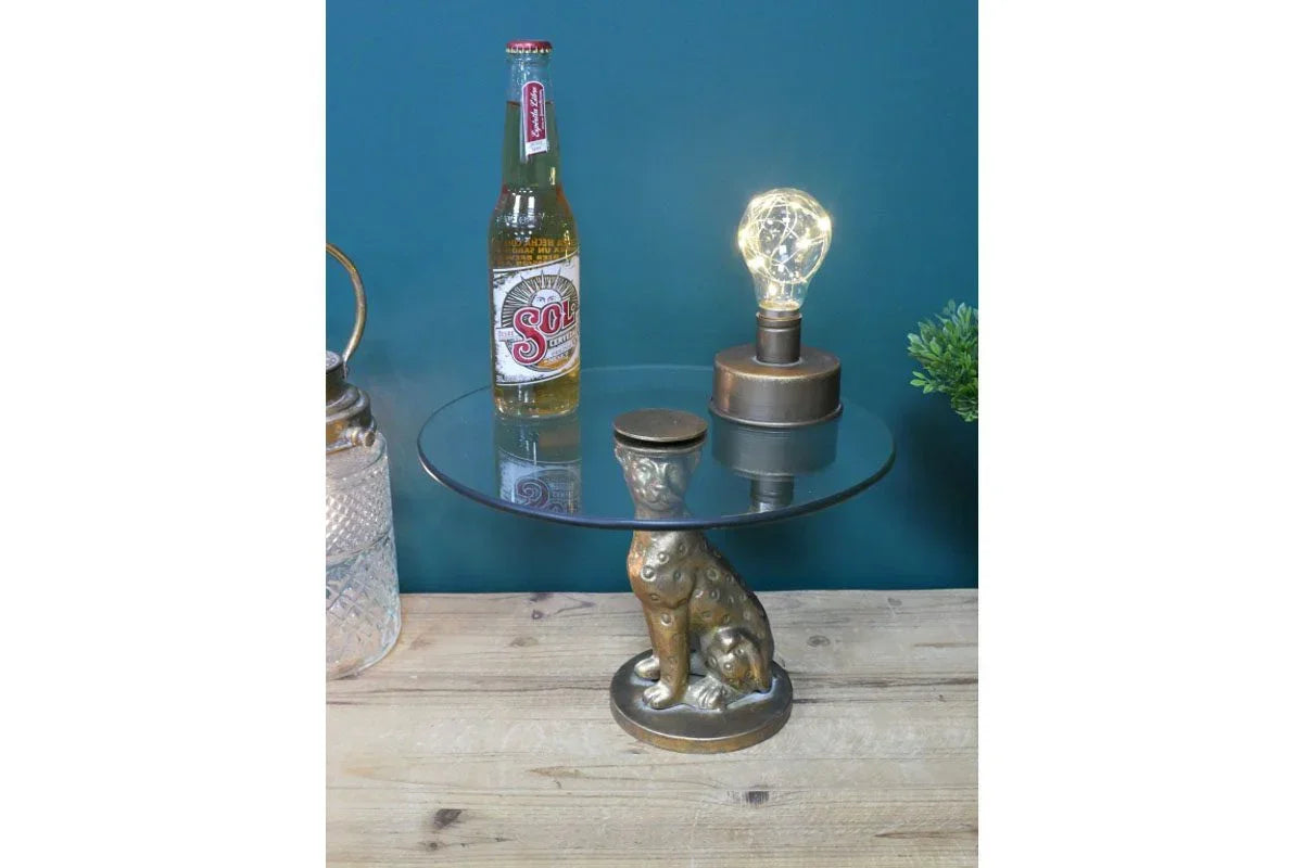 Glass top side table with bronze dog base, lamp and bottle styled on wooden surface