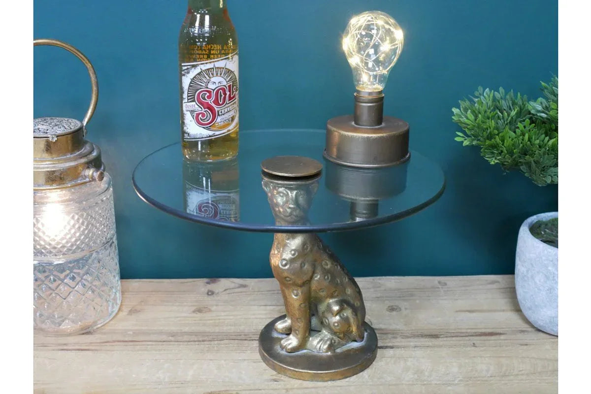 Glass side table with gold leopard base on wooden surface, small lamp and bottle on top