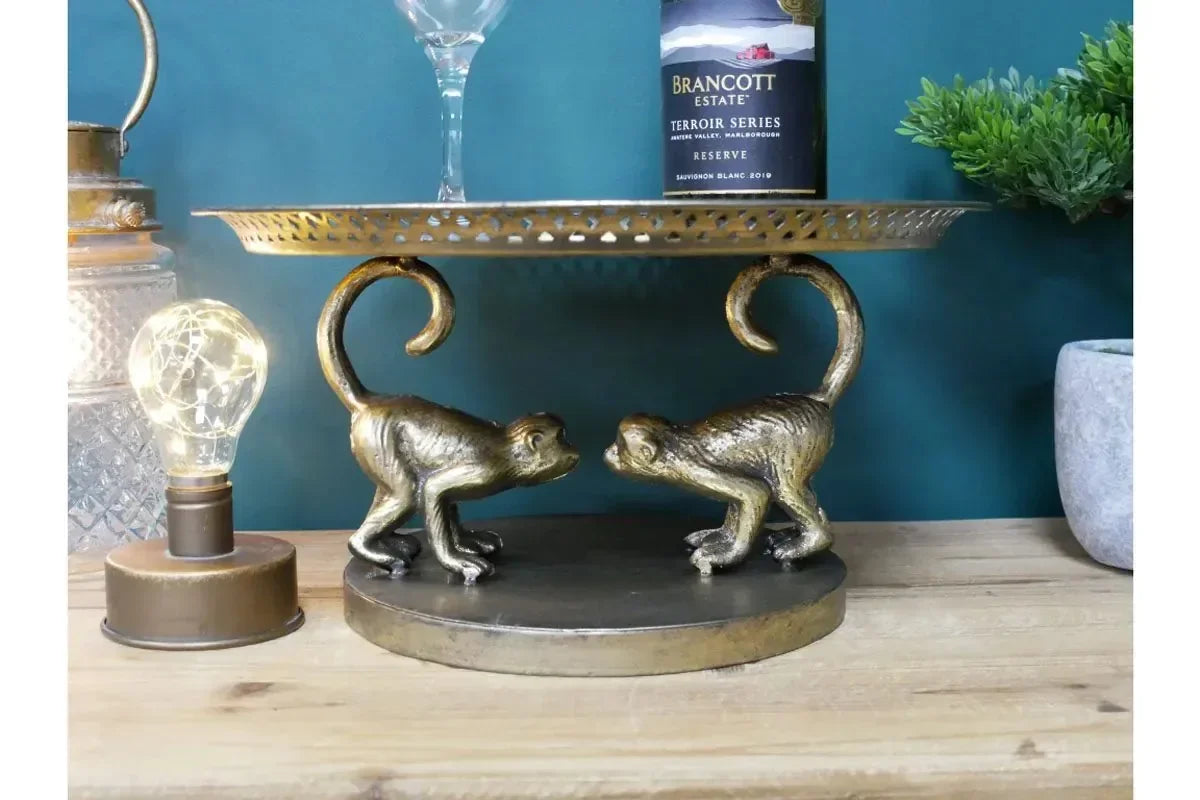 Gold metal cake stand with two monkey figurines, wine bottle, and glass on wooden surface