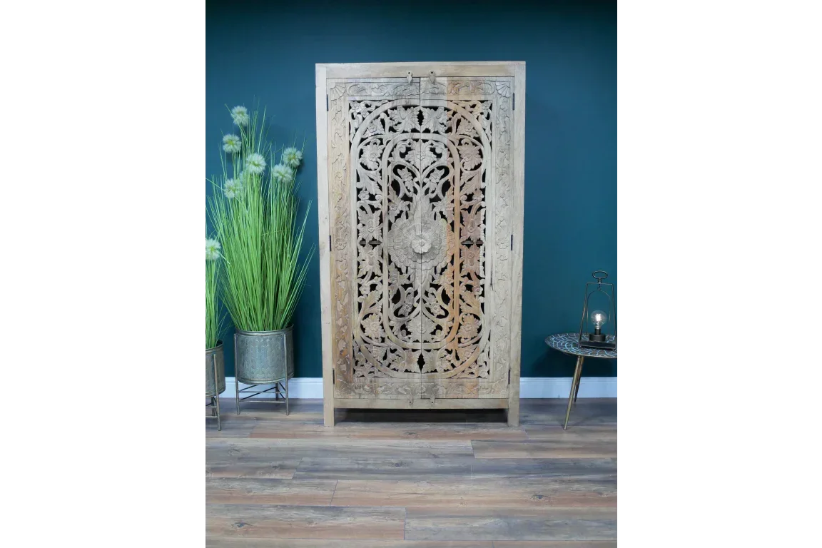 Carved wood wardrobe with floral design against teal wall, next to tall planter and side table