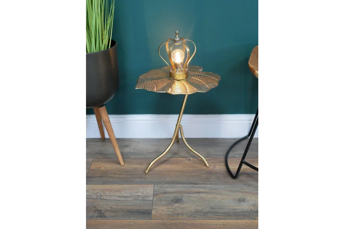 Gold metal side table with leaf design top next to planter and stool on wooden floor