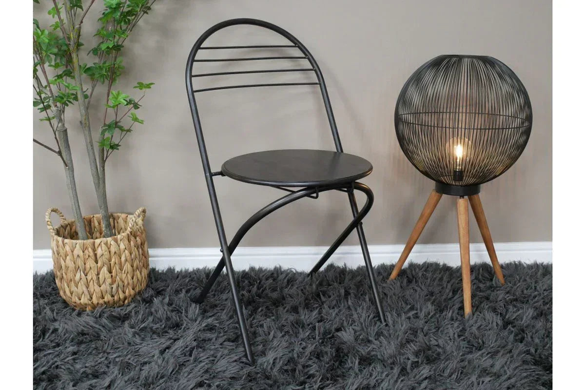 Black metal folding chair on grey rug beside modern tripod floor lamp and woven plant basket