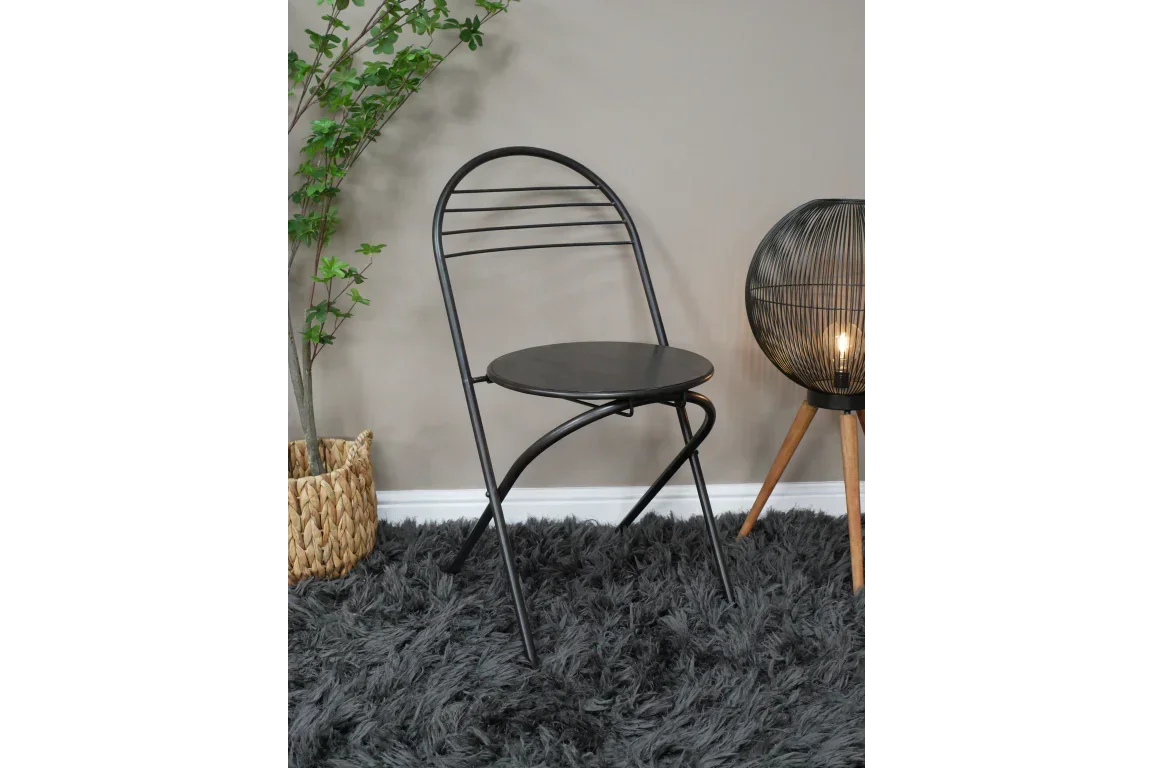 Black metal folding chair with round seat on grey rug beside lamp and indoor plant