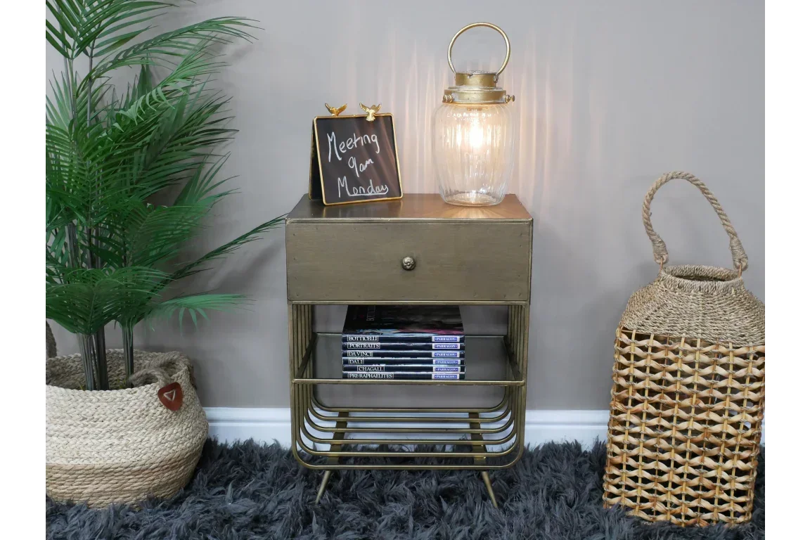 Gold metal bedside table with drawer and shelf, styled with books, lantern, and decorative plant
