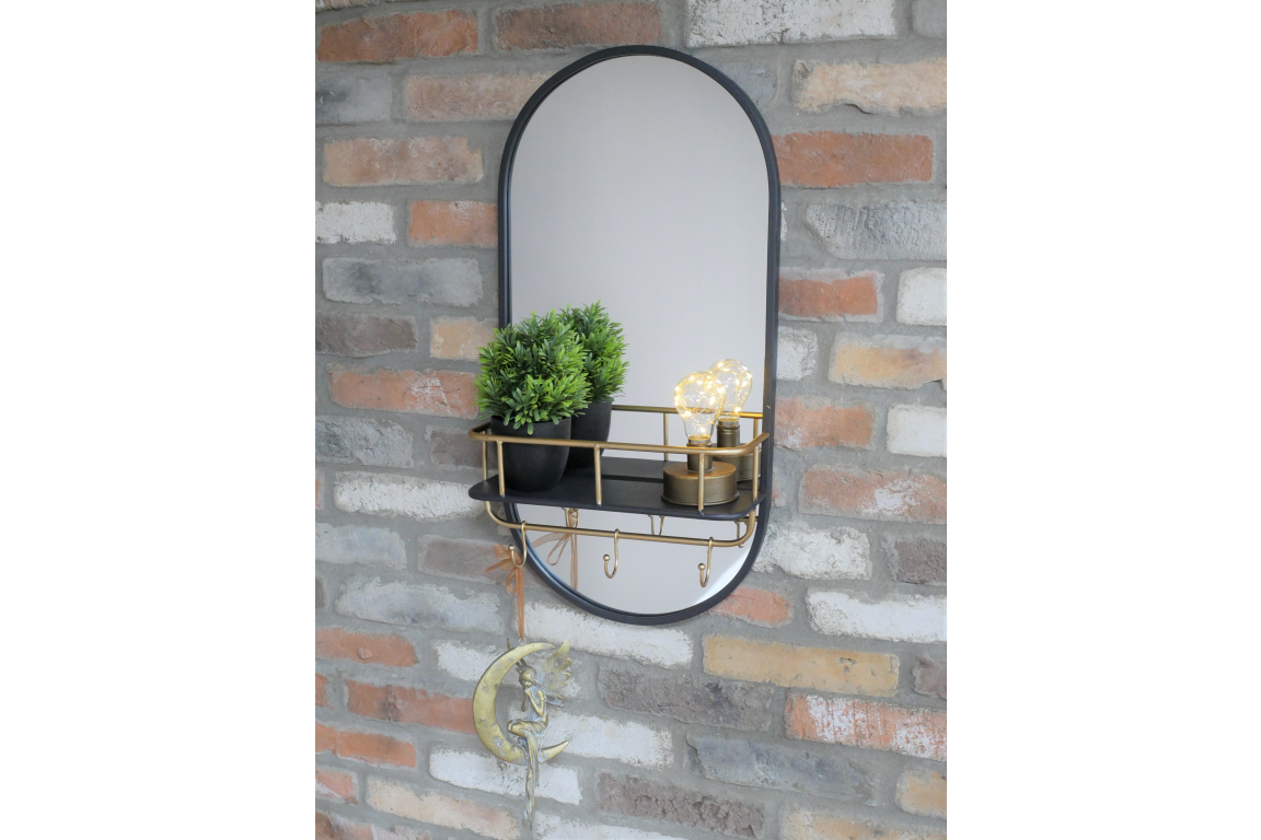 Oval wall-mounted mirror with black frame, gold metal shelf and hooks, shown with potted plant and decorative bulbs.