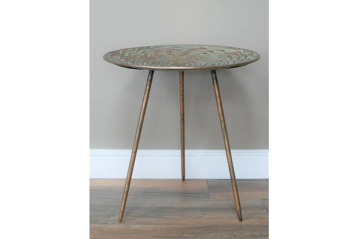 Round metal side table with etched pattern and three slender legs on hardwood floor