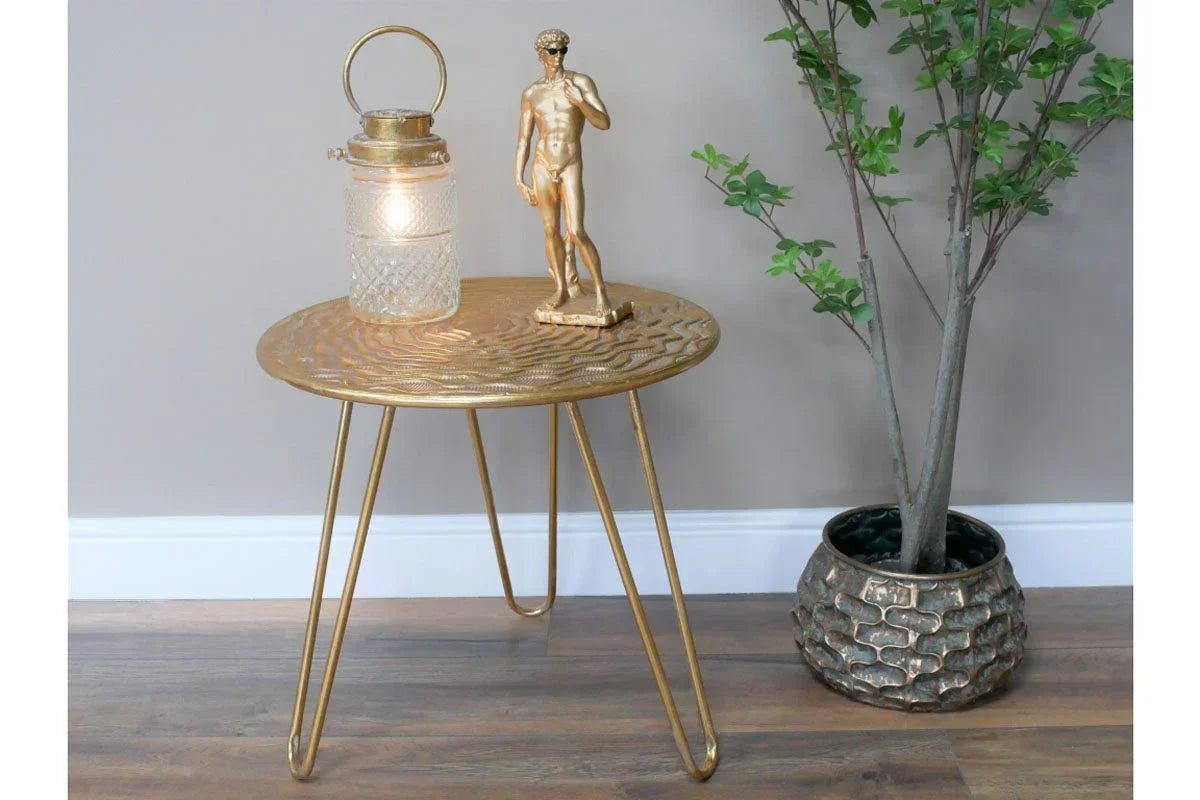 Gold metal side table with textured top holding a lantern and statue, next to faux potted plant