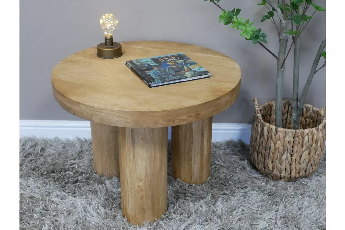 Round wooden coffee table with three thick legs, book and lamp on top, shaggy rug below