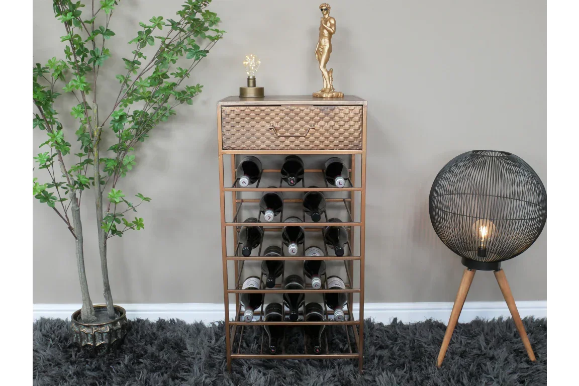 Gold metal wine rack with textured drawer, filled wine bottles, next to floor lamp and plant