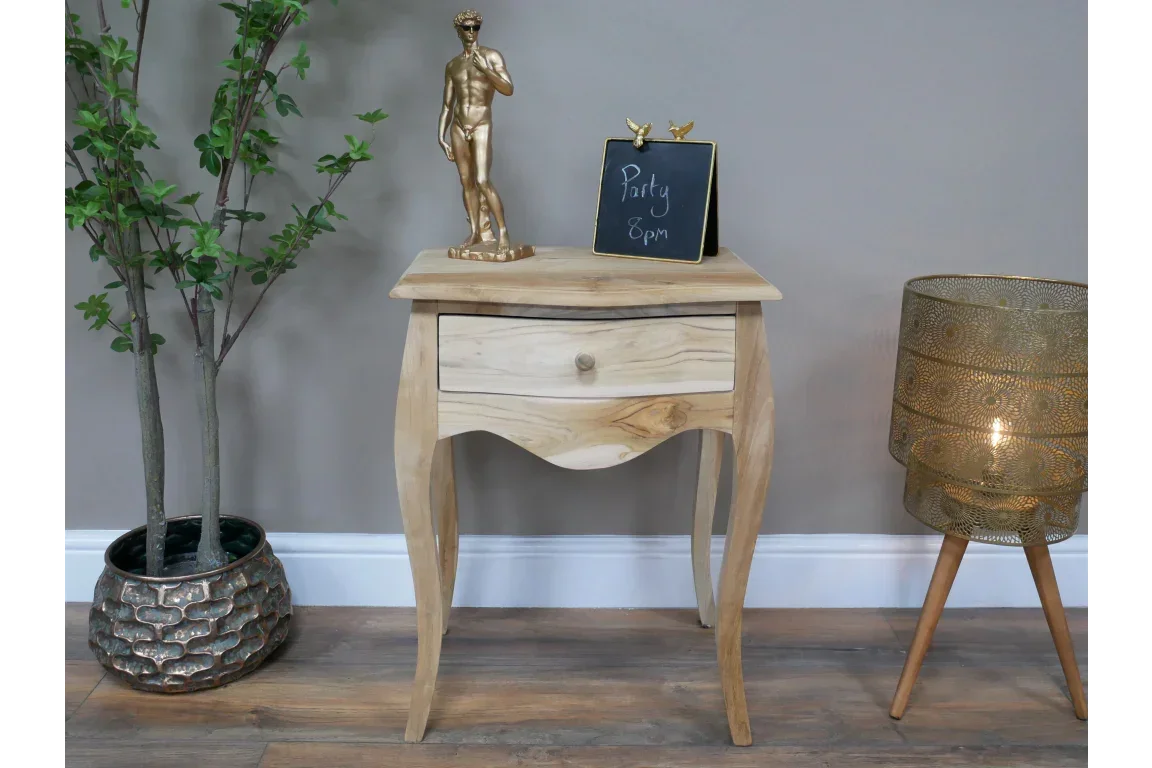 Solid wood bedside table with single drawer, curved legs, decorative lamp and plant nearby