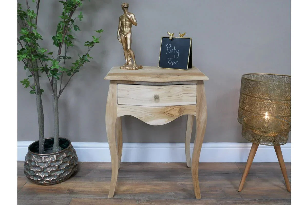 Light wood bedside table with single drawer, decor accents, flanked by plant and gold floor lamp