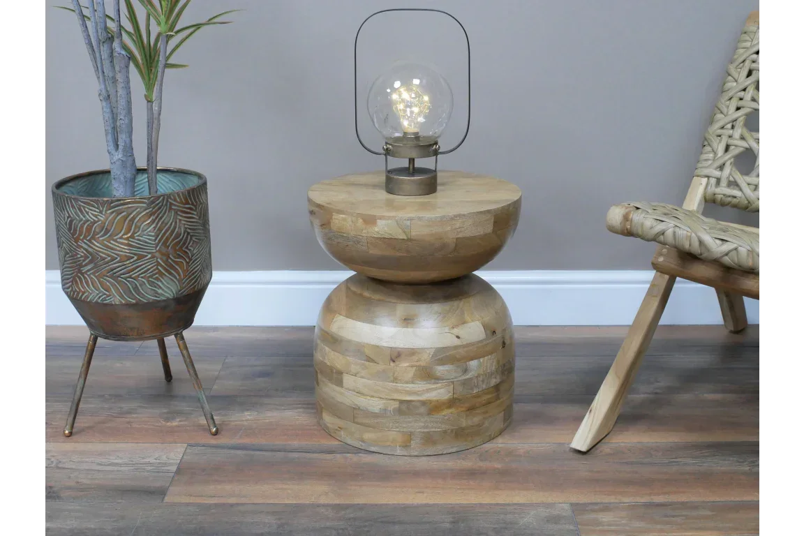 Round wooden side table with natural finish, topped with decorative lamp, beside rattan chair
