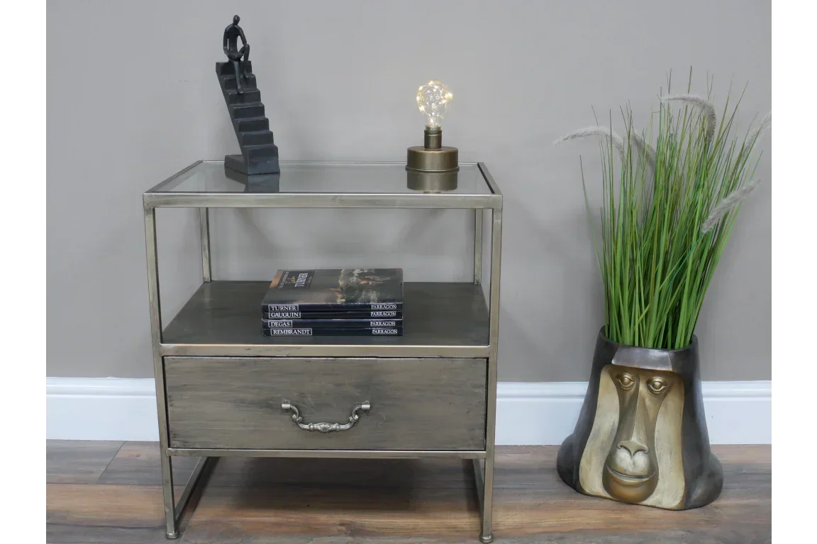 Glass and metal side table with drawer, books, lamp, and sculpture beside decorative plant pot