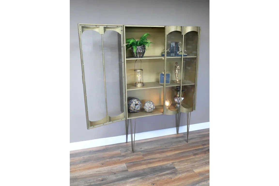 Gold metal display cabinet with glass doors and shelves, standing on tall legs, decor items inside