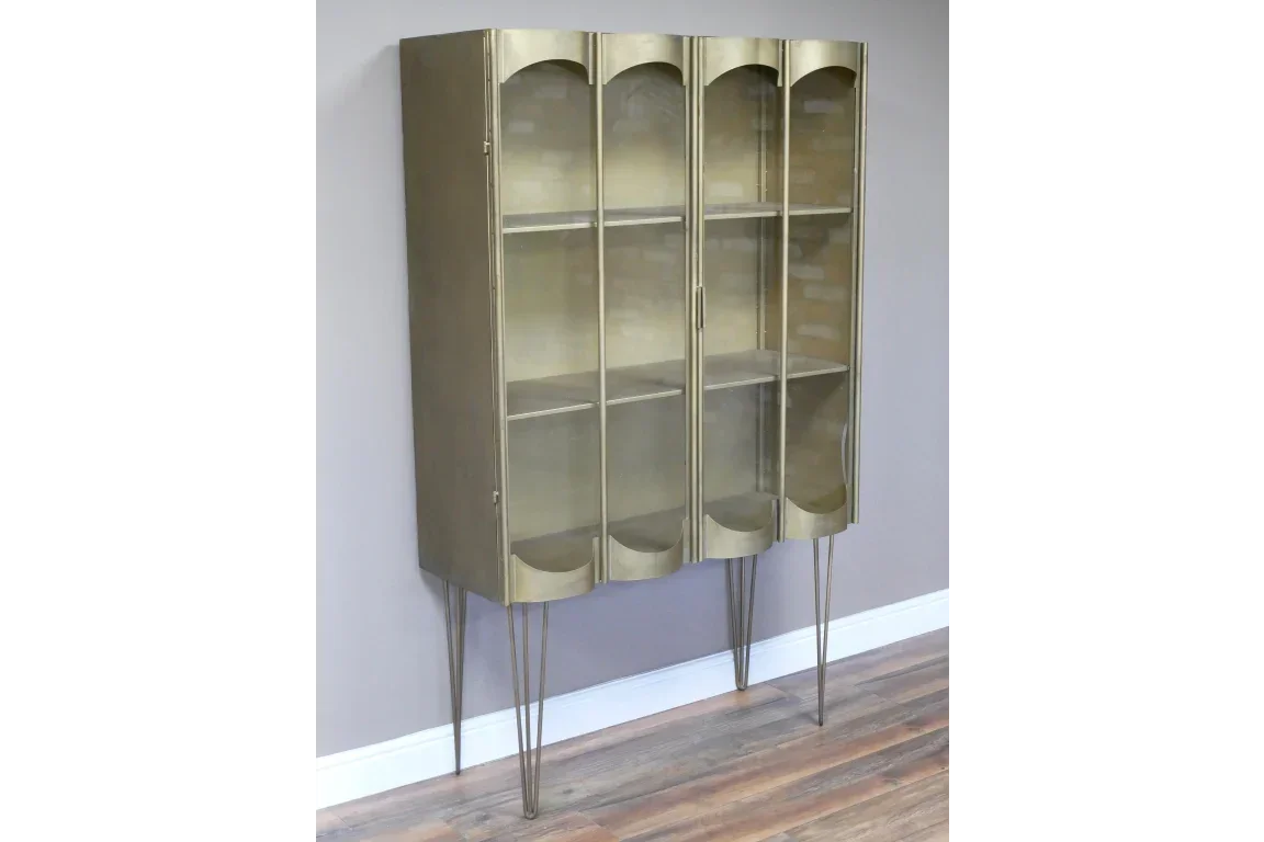 Gold metal display cabinet with glass doors and slim legs against a grey wall