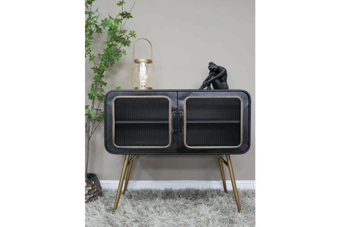 Black sideboard cabinet with gold legs and glass doors, staged on grey rug beside green plant