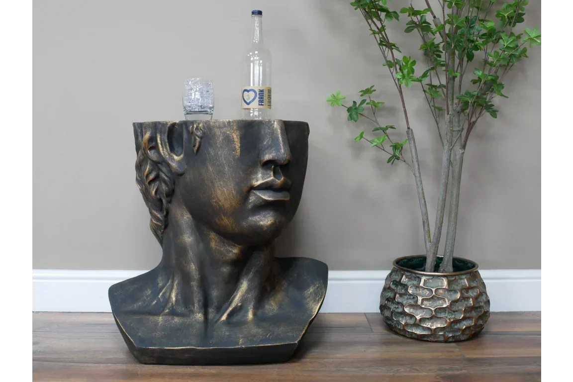 Bronze effect bust side table with flat top, glass and bottle, beside decorative indoor plant