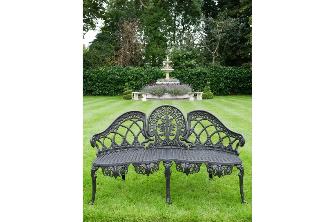 Black cast iron garden bench with ornate backrest on lawn, fountain in background