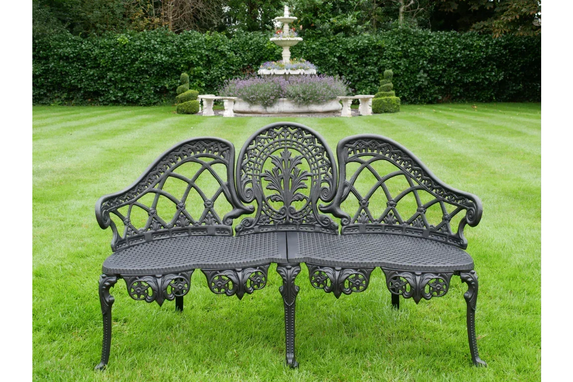 Black cast iron garden bench with ornate backrest on lawn, fountain and greenery behind