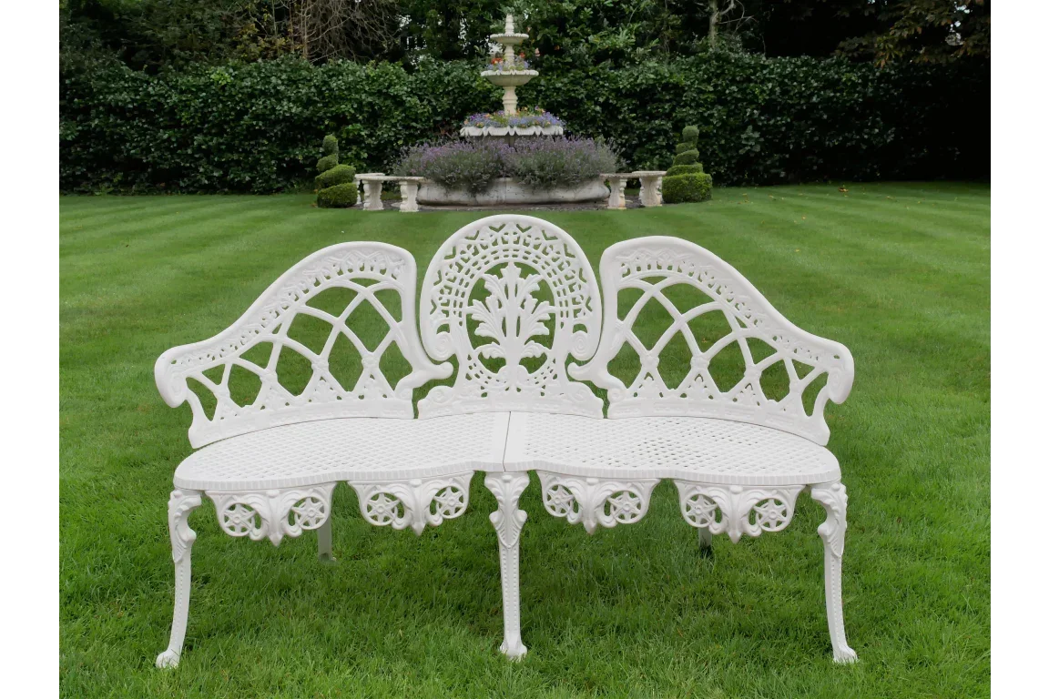 White cast iron garden bench with decorative backrest on lawn, fountain in background
