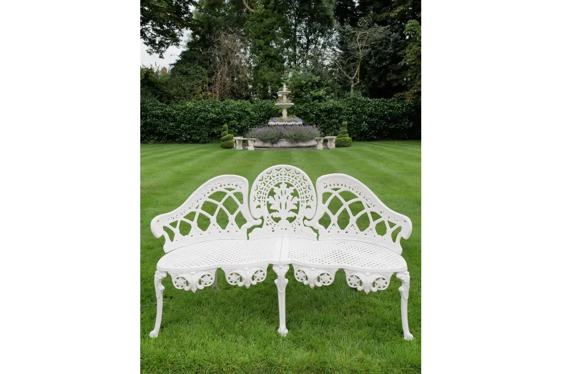 White cast iron garden bench with ornate design on lawn, fountain in background