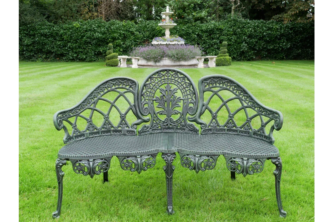 Cast iron garden bench with ornate backrest on lawn, fountain and hedges in background