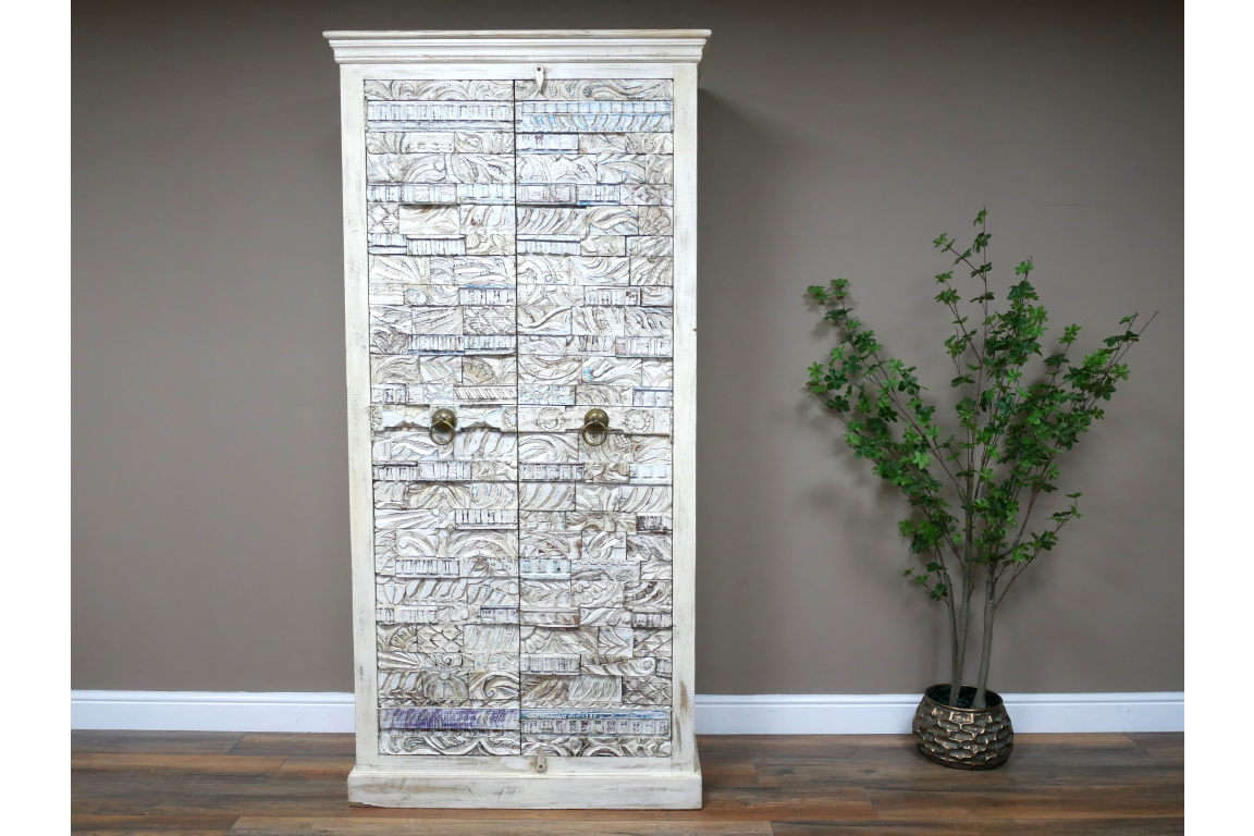 Large white wooden cabinet with intricate carved details and metal ring handles, standing on wooden floor beside green plant.
